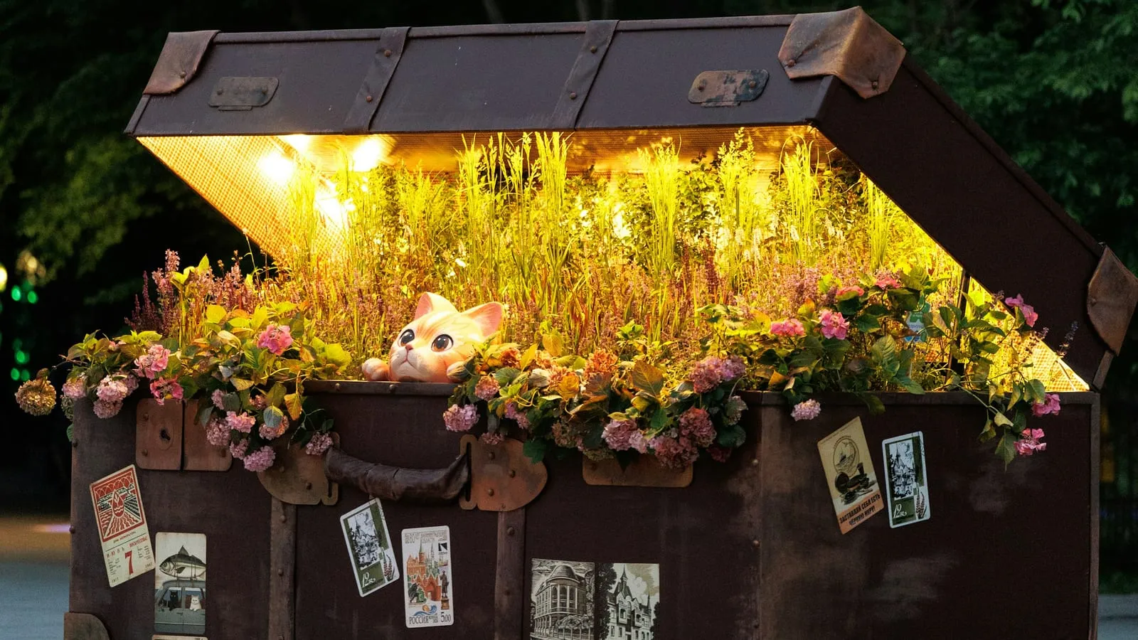 An old steamer trunk at night, lid open and lit from inside, overflowing with grass and pink flowers, a toy cat peeking out.