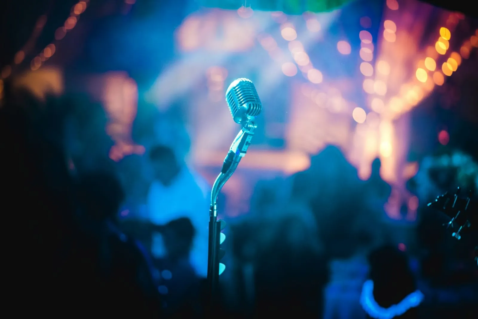 A vintage silver microphone on a stand, in front of a blurred crowd under blue and amber stage lights.